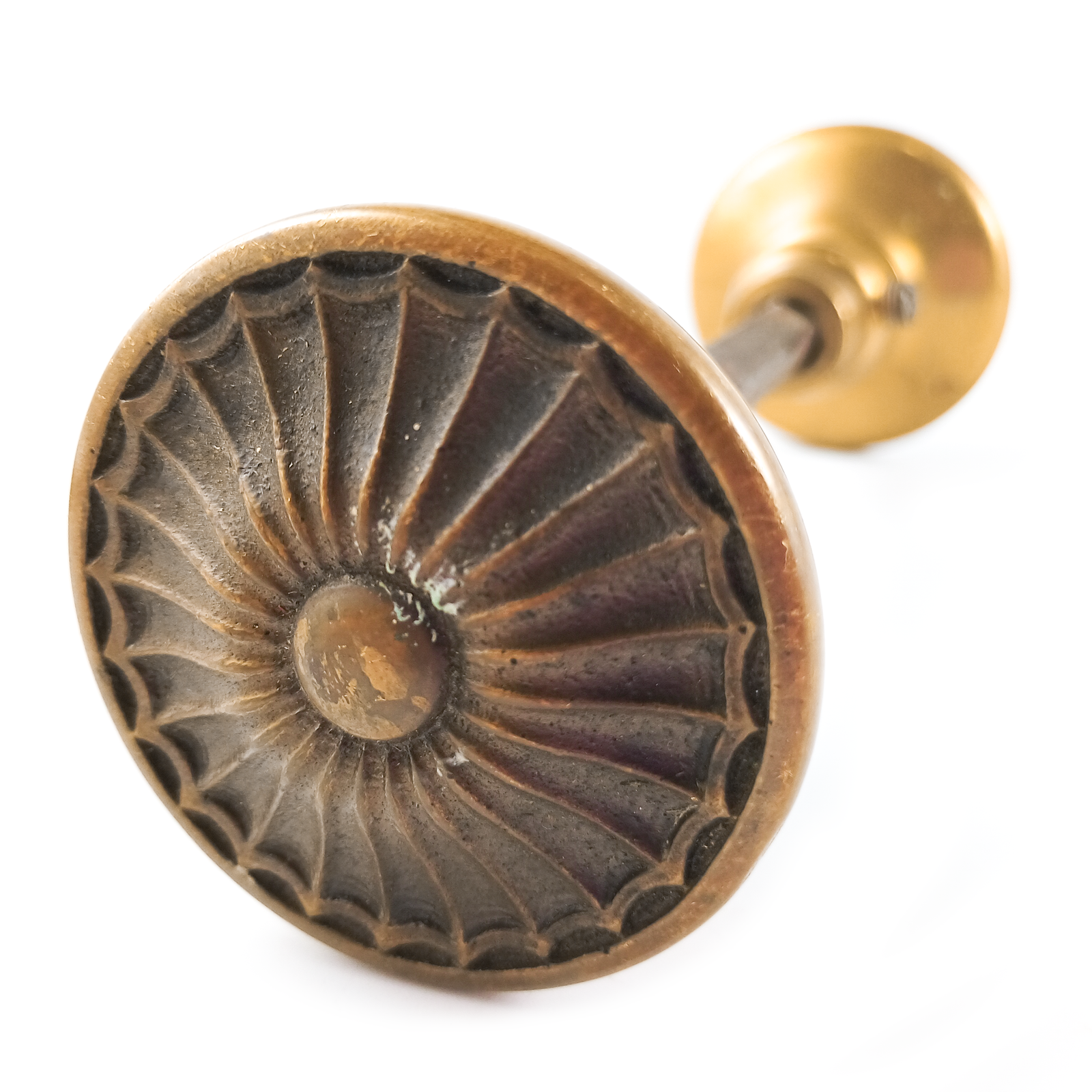 One of the two doorknobs - they have a radial pattern resembling a sunburst, a flower, or a spiderweb around a central round "button," with patina emphasizing the pattern. This knob is a dark, orange-y shade of brass.