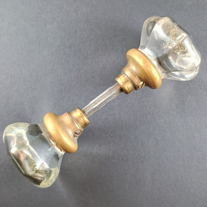 A set of doorknobs threaded onto a spindle, photographed on a grey background. The doorknobs are made of clear glass in a slightly fluted octagonal shape, and have brass bases with some patina on them.