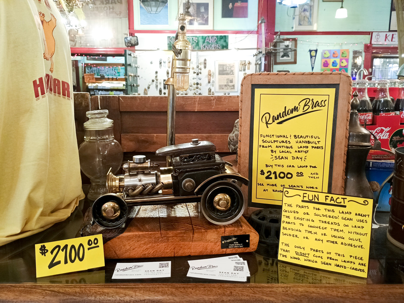A lamp in the shape of an old car, with a price sign stating that it costs $2100. Two signs explain that it was created by a local artist under his company Random Brass, and that it is made of salvaged antique lamp parts, threaded together instead of glued or soldered.