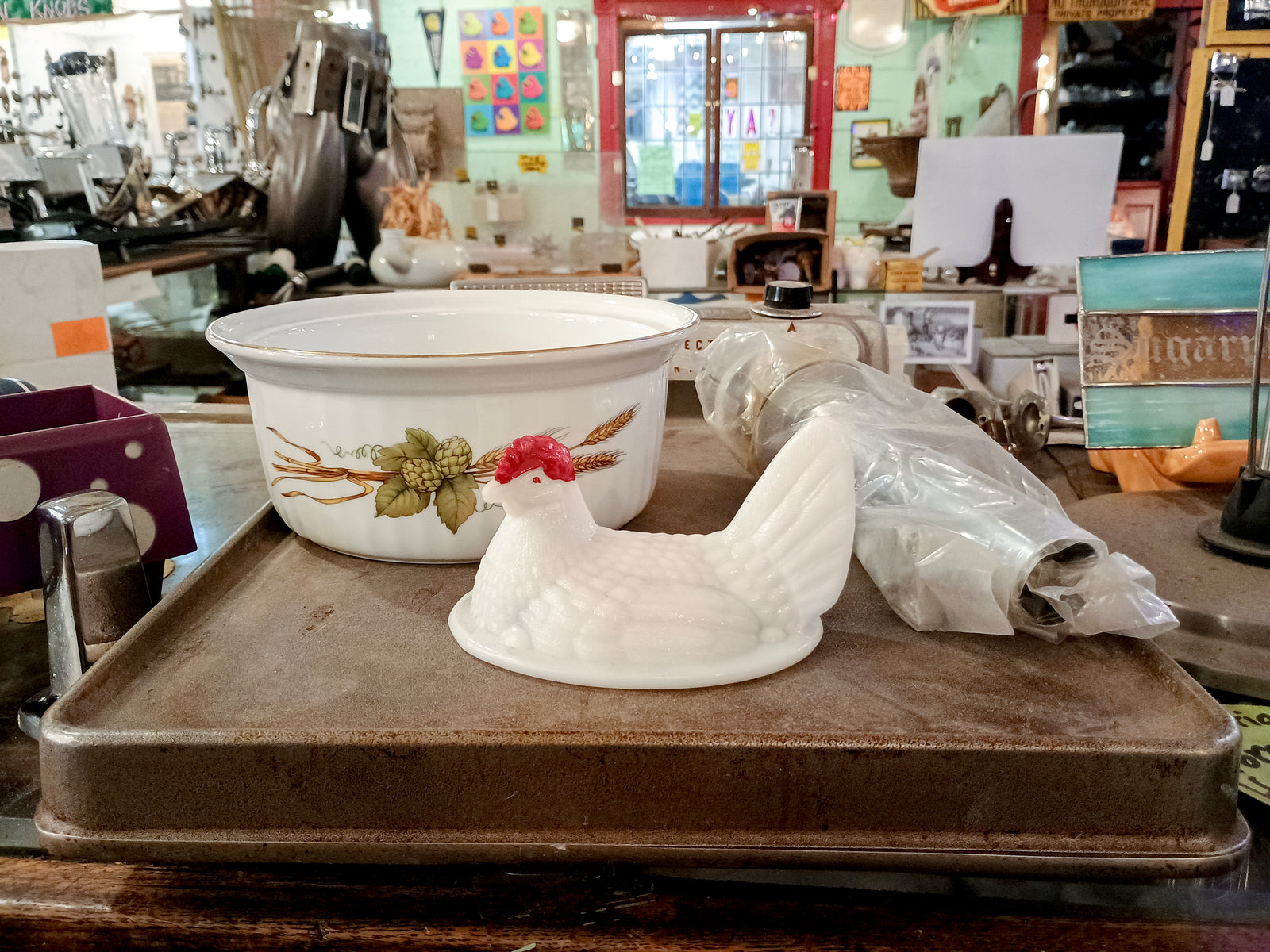 A photo of a ceramic cooking dish with a design consisting of hops and wheat, the top half of a milk glass chicken storage jar, and a plumbing part wrapped in plastic. All three items are sitting on top of an electric griddle, in front of a cluttered background.