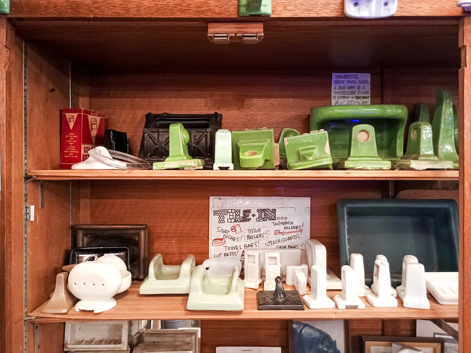 Two shelves of tile-in ceramic bathroom accessories, in varying shades of white, green, blue, and black. There is a sign advertising them as such, as well as various hippos and knick-knacks mixed in.