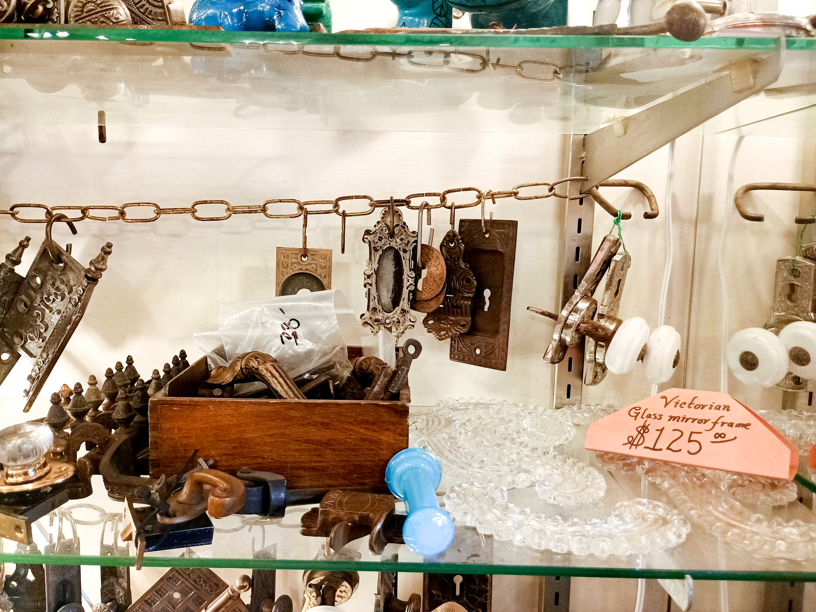 A shelf inside a glass cabinet with several pieces of antique hardware, including some ornate curved pieces of glass with a sign reading "Victorian glass mirror frame: $125"