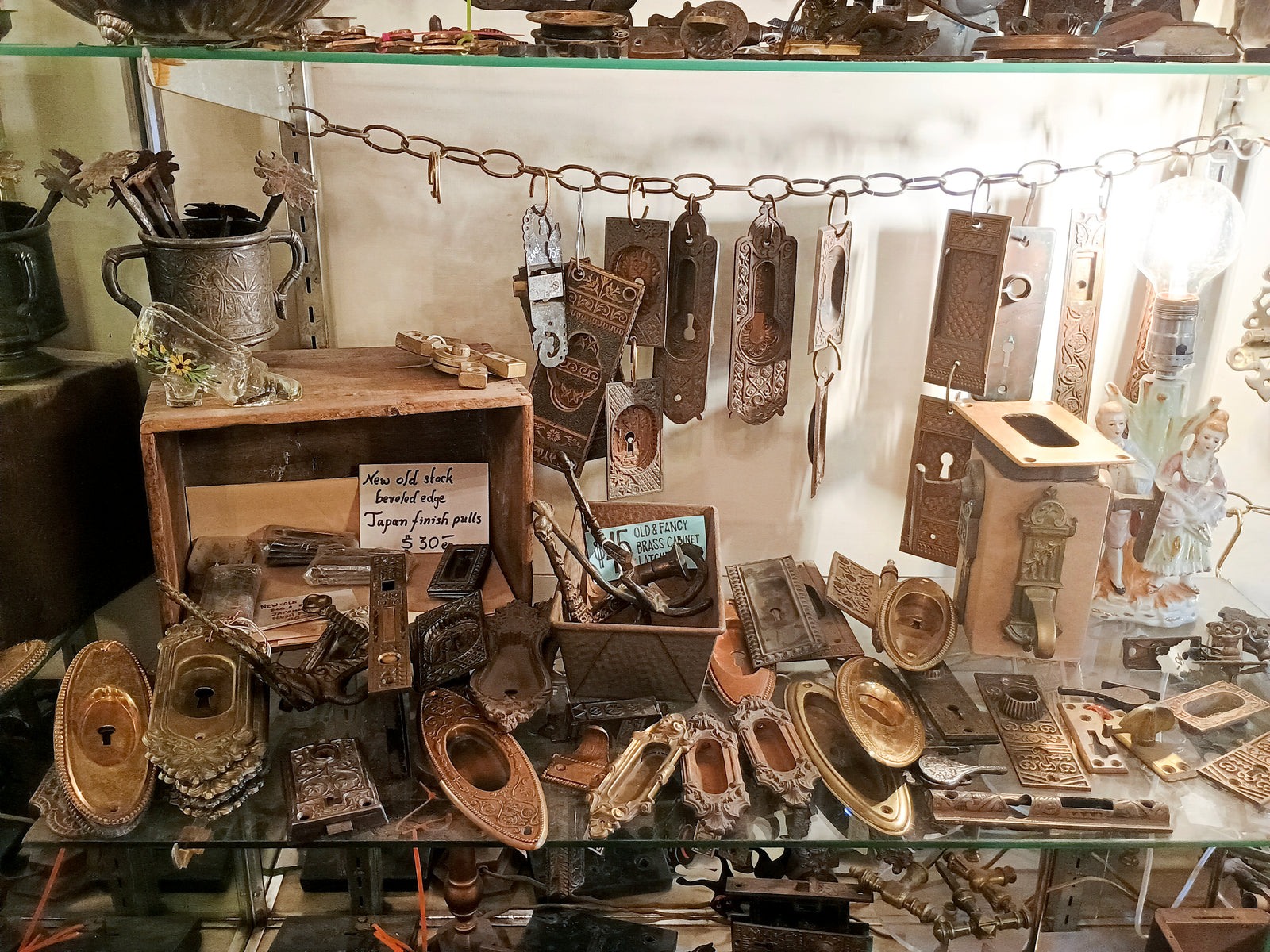 A shelf inside a glass cabinet full of antique pocket door pulls, window lifts, latches, and other various pieces of hardware