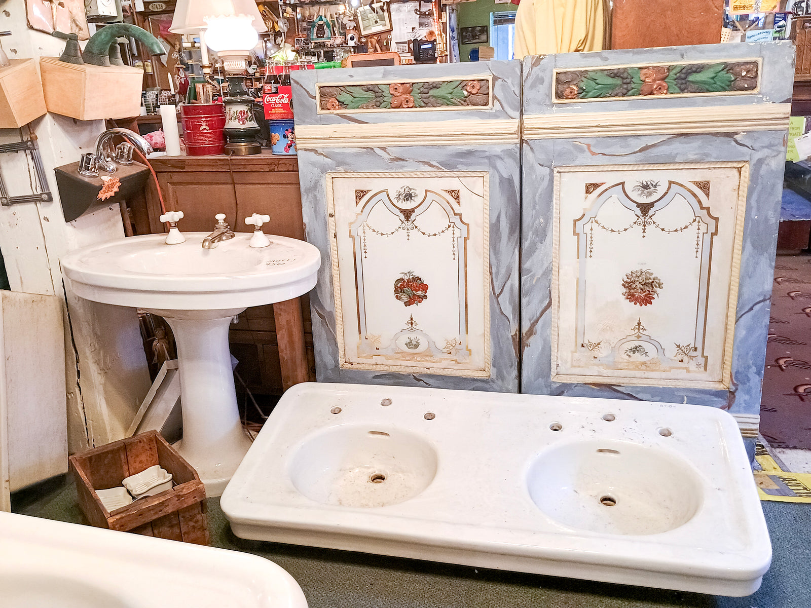A large pedestal sink, a wide double-sink sitting on the ground, and two very large and ornate painted cabinet doors, as well as some various vintage bits and bobs