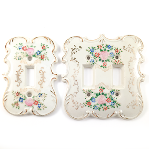 Two porcelain light switch plates, one single-gang, one double-gang. They are both decorated with hand-painted floral designs in pink, red, and blue, with green leaves, as well as with swirling gilded designs. The edges of each plate are ornately curved and raised.
