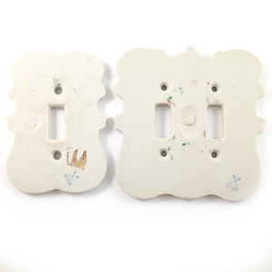 The backs of both light switch plates, which have some paint staining on them, as well as stamps. The single-gang plate also has the illegible remnants of a sticker on it.