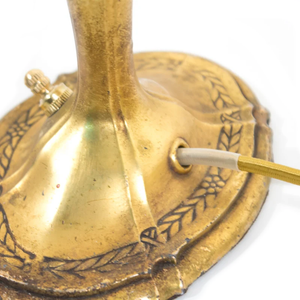 The base of an Art Nouveau brass table lamp shaped like a candlestick, showing the ornate floral detailing and the cord coming out of the base