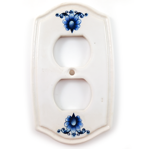 A white porcelain double outlet cover, with very slightly curved sides and a more significantly curved top and bottom. At the top and bottom, nestled into the curves, are blue delft flower designs. There is also an indented decorative lip around the edge of the plate.