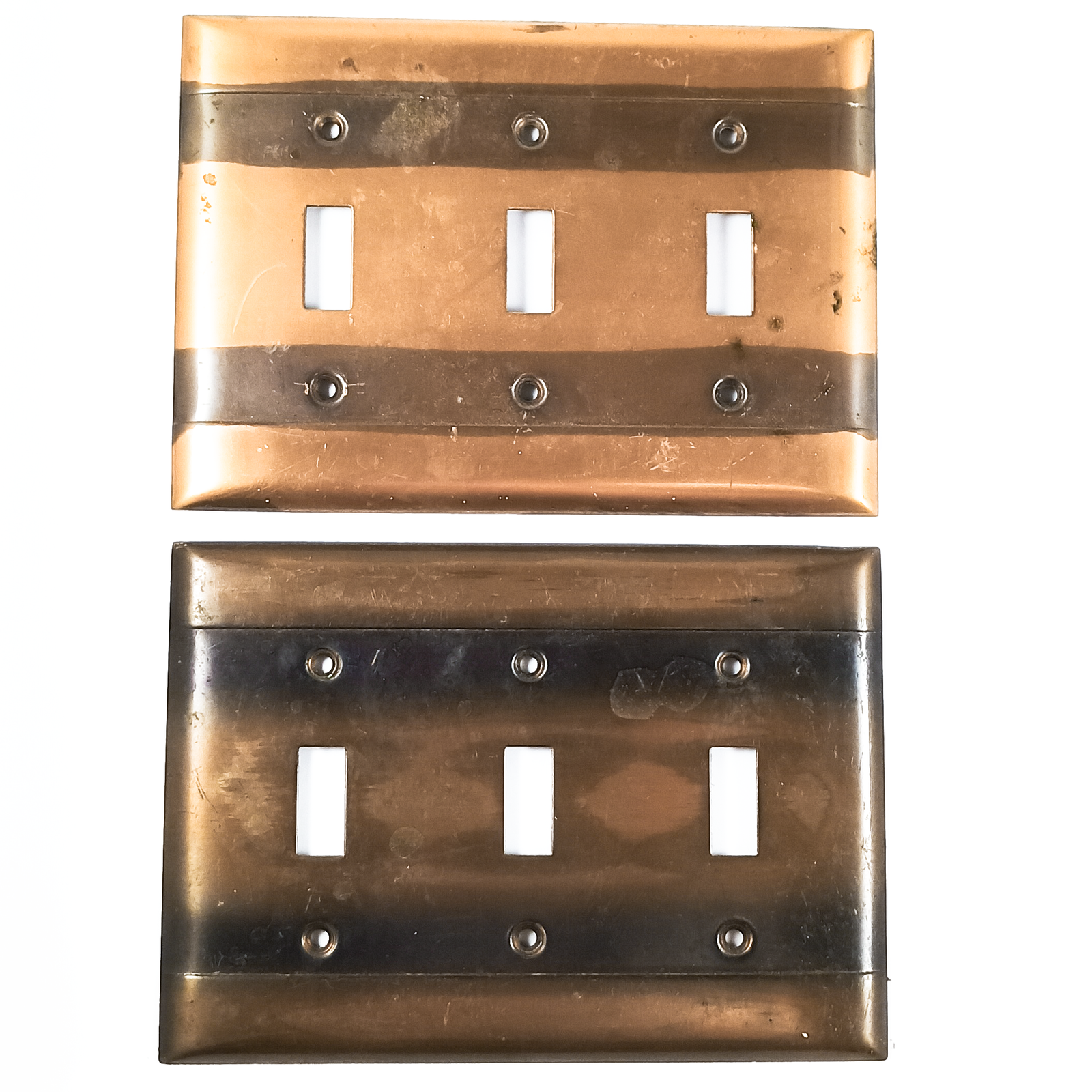 Two light switch plates, each with six screw holes and three openings for switches. One plate is a light bronze, with two very distinct stripes of dark patina going horizontally across the screw holes. The other plate has the same stripes, but is overall darker in finish, and the stripes are blurred at the edges. Both plates have some surface stains or what may be adhesive residue. as well as areas of uneven finish.