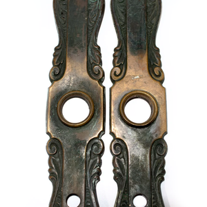 A close-up of two bronze Art Nouveau escutcheons, showing the dark patina and the bronze finish as well as the ornate details