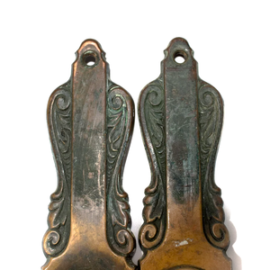 A close-up of the tops of two bronze Art Nouveau escutcheons, showing the ornate details and dark patina