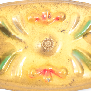 A close-up of the center of a gold-tone vintage ceiling light fixture, showing some of the surface wear and the scrolling raised decorations which have been painted green and red