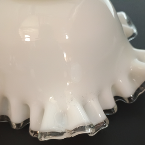 A close-up of damage to the edge of a ruffled milk glass lamp shade with clear glass along the edge. There is some chipping and a crack about one inch long, as well as a very small amount of dark soot-like material