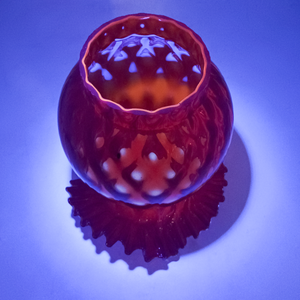 The fitter of a red lamp shade with a ruffled edge, shown under UV light which makes the background appear purple and the inside of the fitter and interior of the shade fluoresce bright orange