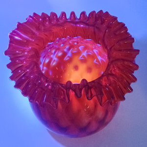 A red lamp shade with a ruffled edge under UV light, making the background appear purplish blue and making the interior of the shade fluoresce orange, along with the very edge of the ruffled rim