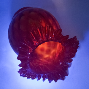 A bright red lamp shade with a ruffled edge, shown under UV light, which makes the background appear purple and the interior of the lamp shade fluoresce bright orange