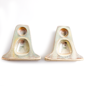 A pair of ceramic wall-mounted toilet paper roll holders. They each have one circular recess and, behind it, one irregular, decorative rounded oval recess. Each piece has a glaze that appears to be a mottled, light green-brown color, with darker brown edges. There is a bright silver sheen to the glaze.