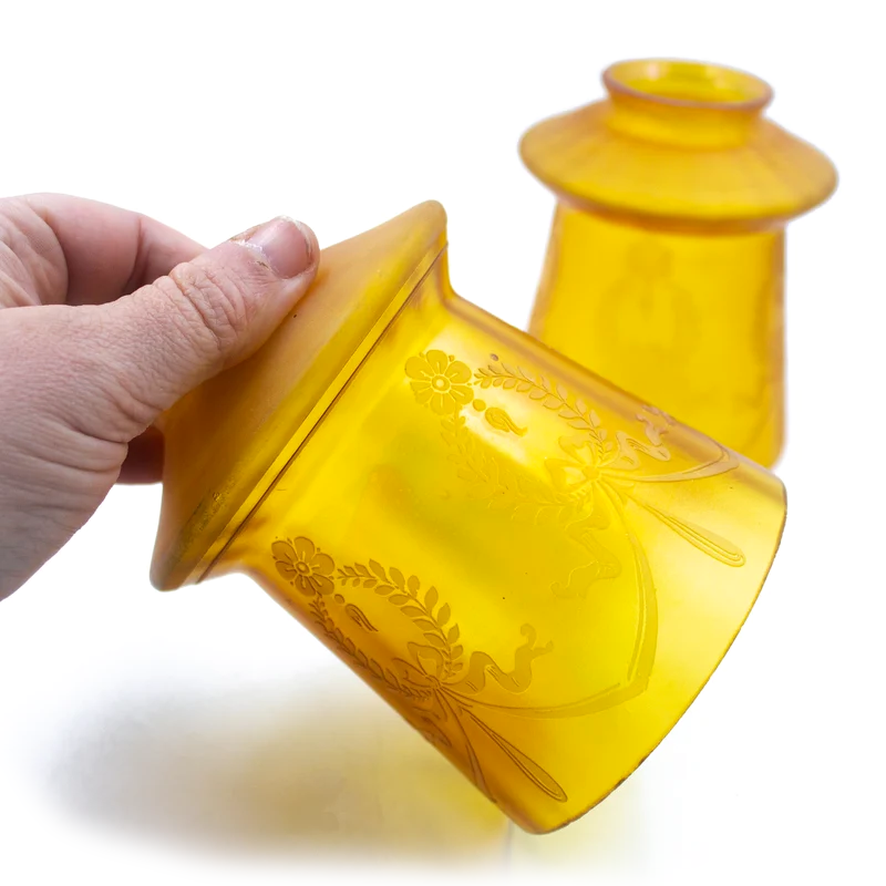 Two yellow-amber glass lamp shades with etched Art Nouveau designs in the frosted finish. One is standing upright, one is on its side, and both show very subtle iridescence on the surface of the glass