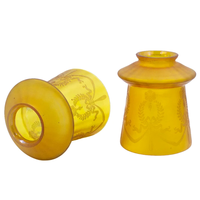 Two yellow-amber glass lamp shades with etched Art Nouveau designs in the frosted finish. One is standing upright, one is on its side, and both show very subtle iridescence on the surface of the glass