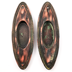Two oval pocket door pulls with a beaded trim and copper-and-black japanned finish. They have some verdigris on the surface, and no key holes.