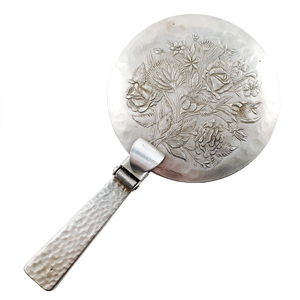 A silver-tone silent butler, which looks somewhat like a pan, with a round portion and a slightly tapered rectangular handle. The round portion is made of hammered aluminum with much of the texture burnished out, and has a very detailed floral design etched into it. The handle is also hammered aluminum, but with a much more distinct texture. There is a curled piece of brushed aluminum where the two pieces meet, which is used to open the lid.