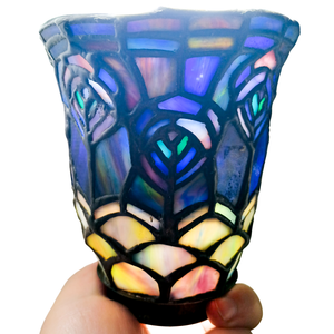 A stained glass lampshade held in a person's hand and illuminated by a light source within. The upper portion of the shade is a pattern resembling peacock feathers, in vivid blues, pinks, purples, and aquas, while the lower portion is a scaled pattern in swirled pinks and yellows.
