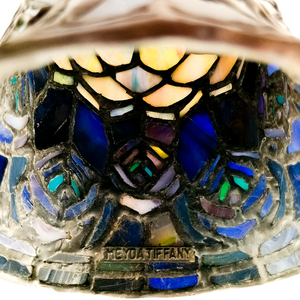 The inside of a stained glass lampshade, partials illuminated to reveal vivid shades of green, aqua, yellow, and blue in the peacock feather pattern. A stamped cartouche near the rim of the shade reads "MEYDA TIFFANY"