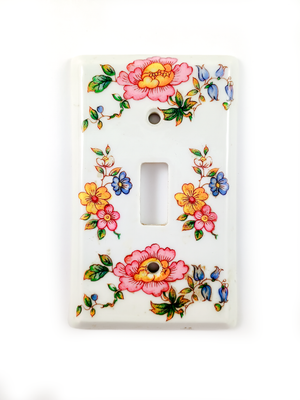 A white, rectangular, single-gang light switch decorated with pink, blue, and yellow flower designs. A small stamp is barely visible on the bottom edge.