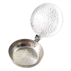 A view of the inside with the lid open, showing a more heavily hammered texture on the inside. The hammered texture has been worn down smooth around the edge, likely from contact with the bowl. The bowl is also hammered aluminum, and is riveted to the handle with a triangle of three large rivets, while the lid is riveted on with two. The Canterbury logo - a knight's helmet in profile - is stamped into the underside of the lid on the edge, though it is partly cut off by the edge of the lid.