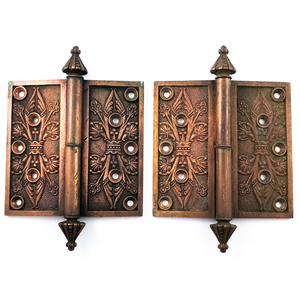 A pair of complete hinges with ornate, abstracted floral patterns on the faces. They have cone-shaped, decorate finials, and five screw holes in each face. There is significant patina on each hinge piece, and a small amount of verdigris, but the details are very well-preserved.
