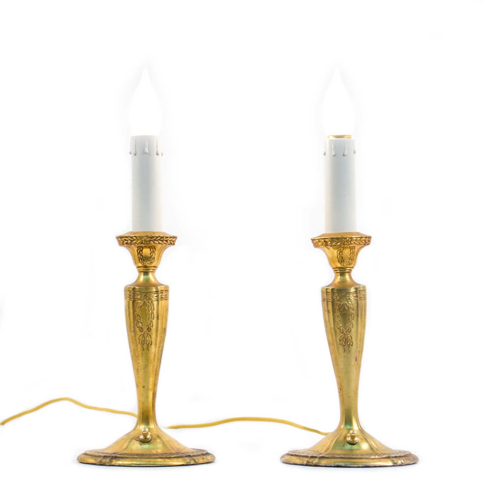 A pair of brass table lamps shaped like candlesticks, complete with faux candles. They have flame-shaped bulbs installed for display, and the brass bases have ornate flower and leaf details.