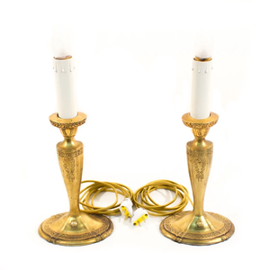 A pair of brass table lamps shaped like candlesticks, complete with faux candles. They have flame-shaped bulbs installed for display, yellow vintage-styled cords, and the brass bases have ornate flower and leaf details.
