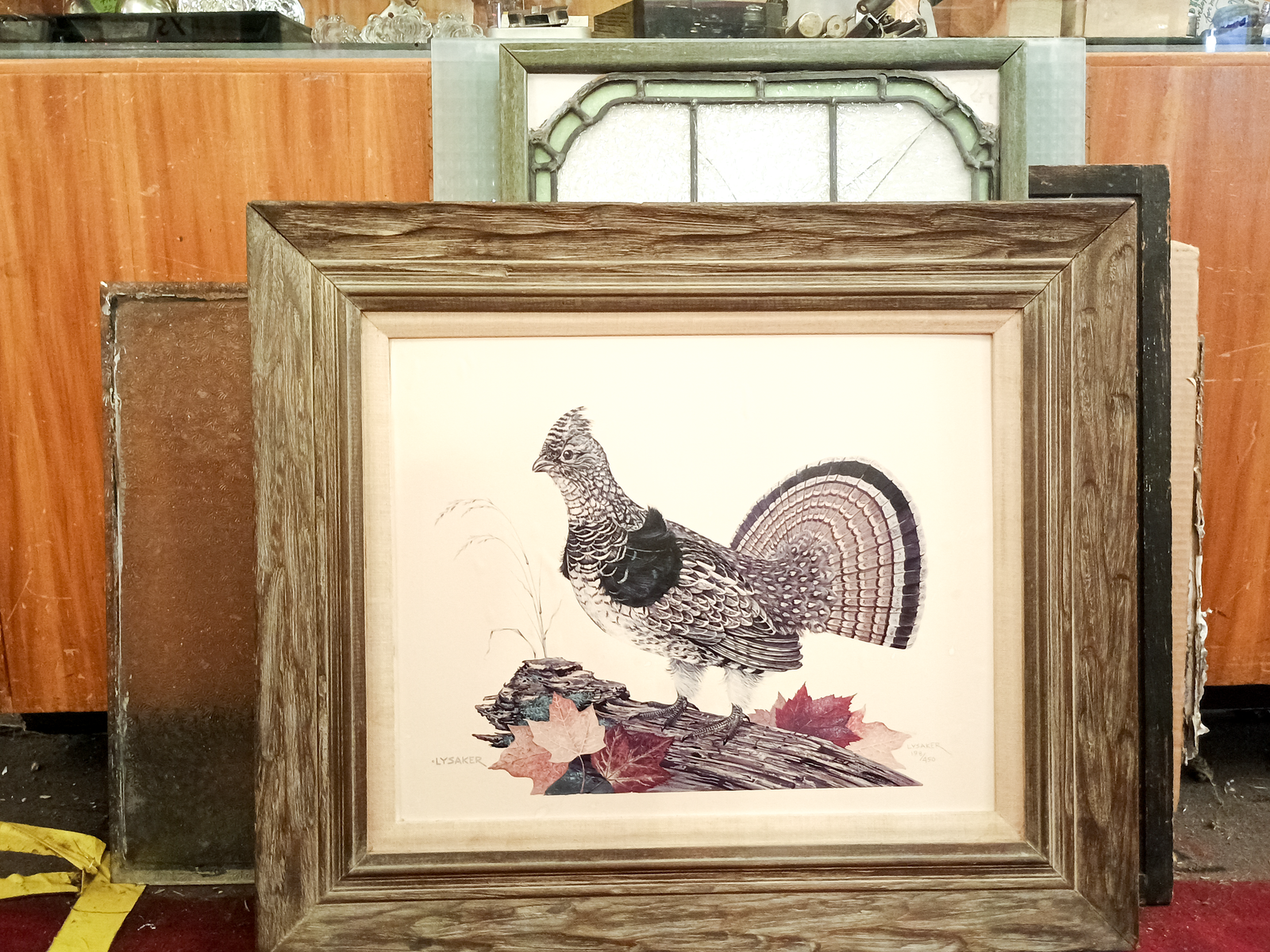 A photo of a print inside a large wooden frame. The print is of a pheasant standing on a log with fallen leaves around it, and says "LYSAKER" in one corner. There is a stained glass window and a pane of glass partially visible behind the framed print.