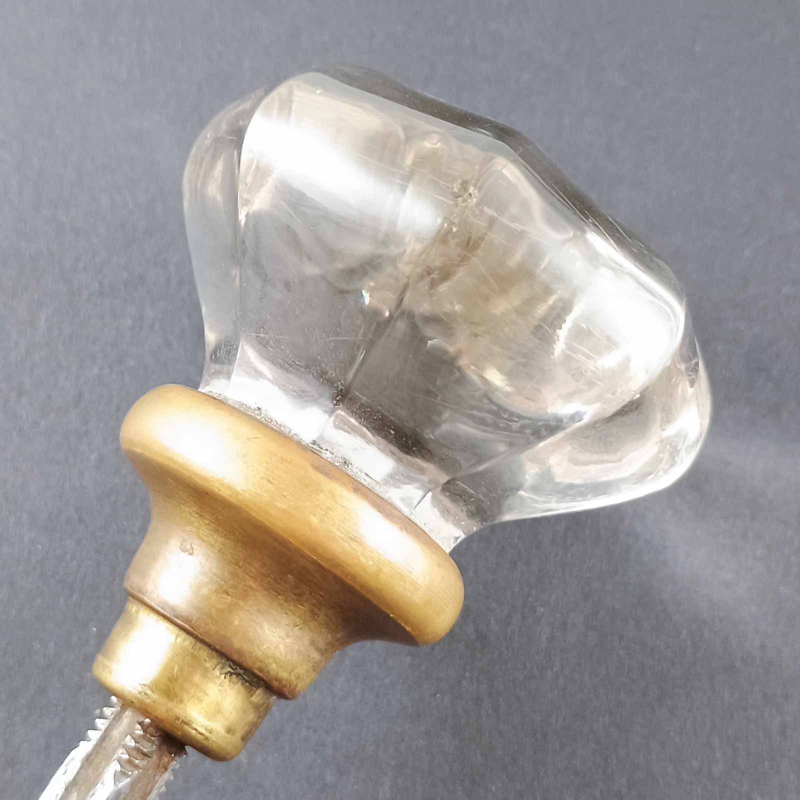 A set of doorknobs threaded onto a spindle, photographed on a grey background. The doorknobs are made of clear glass in a slightly fluted octagonal shape, and have brass bases with some patina on them.