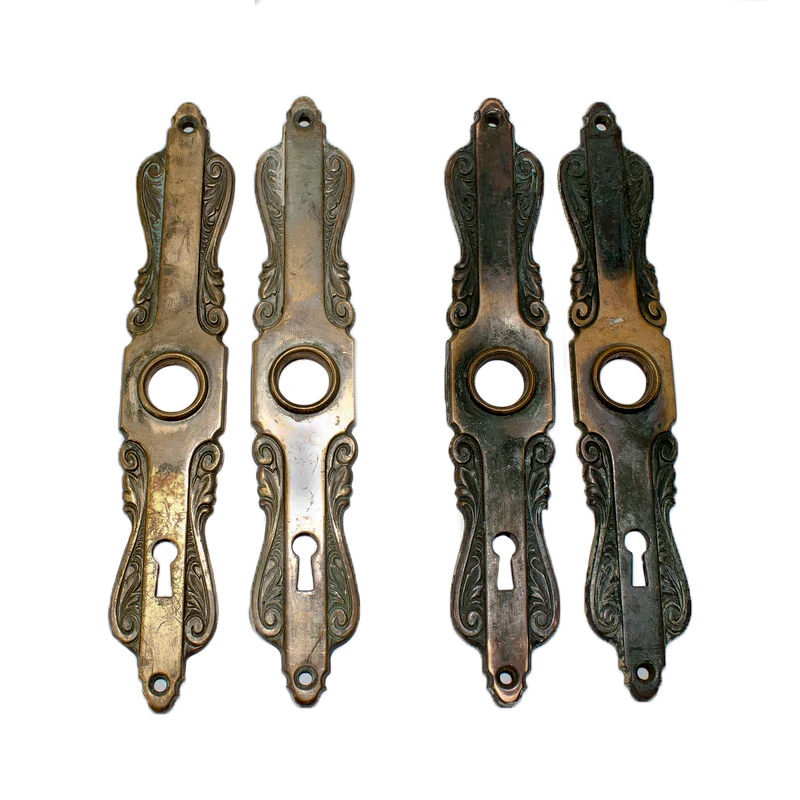 Two sets of Art Nouveau escutcheons, with intricate scrolling details, a central bezel for a doorknob, and keyholes at the bottom. Two of the escutcheons have a lighter brass finish, and two have a dark patina-coated bronze finish.