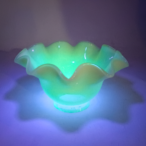 The lamp shade upright on its base under UV light, causing the background to appear purple and the shade to fluoresce bright green. The clear rim is more visible here, as it does not fluoresce.