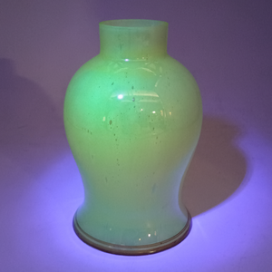 The shade standing on its rim, under UV light. The background appears purple from the light, and the shade is fluorescing bright green. The bubbles, which were previously somewhat difficult to see, stand out more as they do not fluoresce.