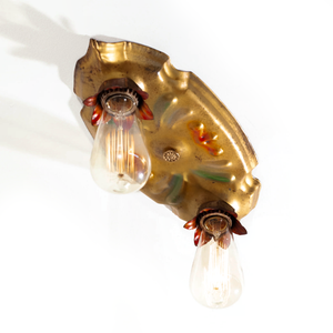 A photo of a vintage gold-tone ceiling light fixture with two sockets, each of which is housed in a red flower shape. There are red and green scrolling decorations in the middle of the fixture, and bulbs have been installed in each socket.