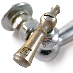 A close-up of a chrome-plated brass lever handle on an antique farmhouse faucet, labeled "H" for hot, which is missing most of its chrome plating